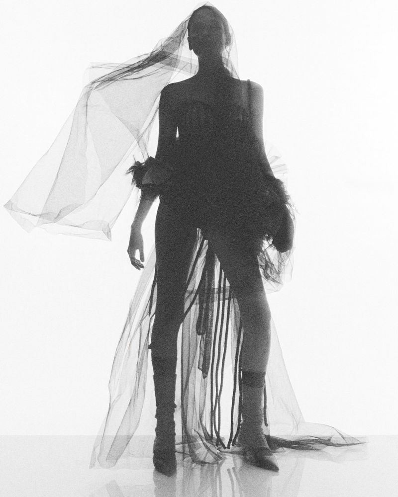 Woman posing in a black dress and veil against a bright white background, creating a high-contrast, fashion-forward silhouette.