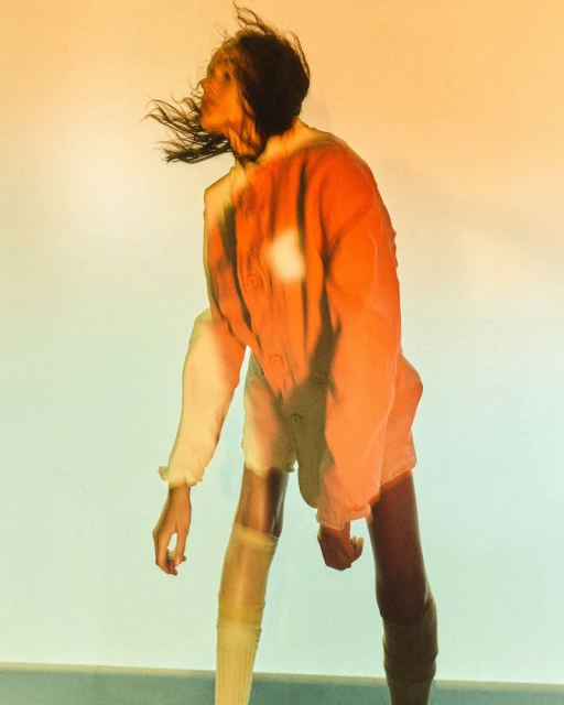 Stylish woman in an orange jacket and white shorts poses indoors against a pale yellow backdrop in a brightly illuminated photo studio.