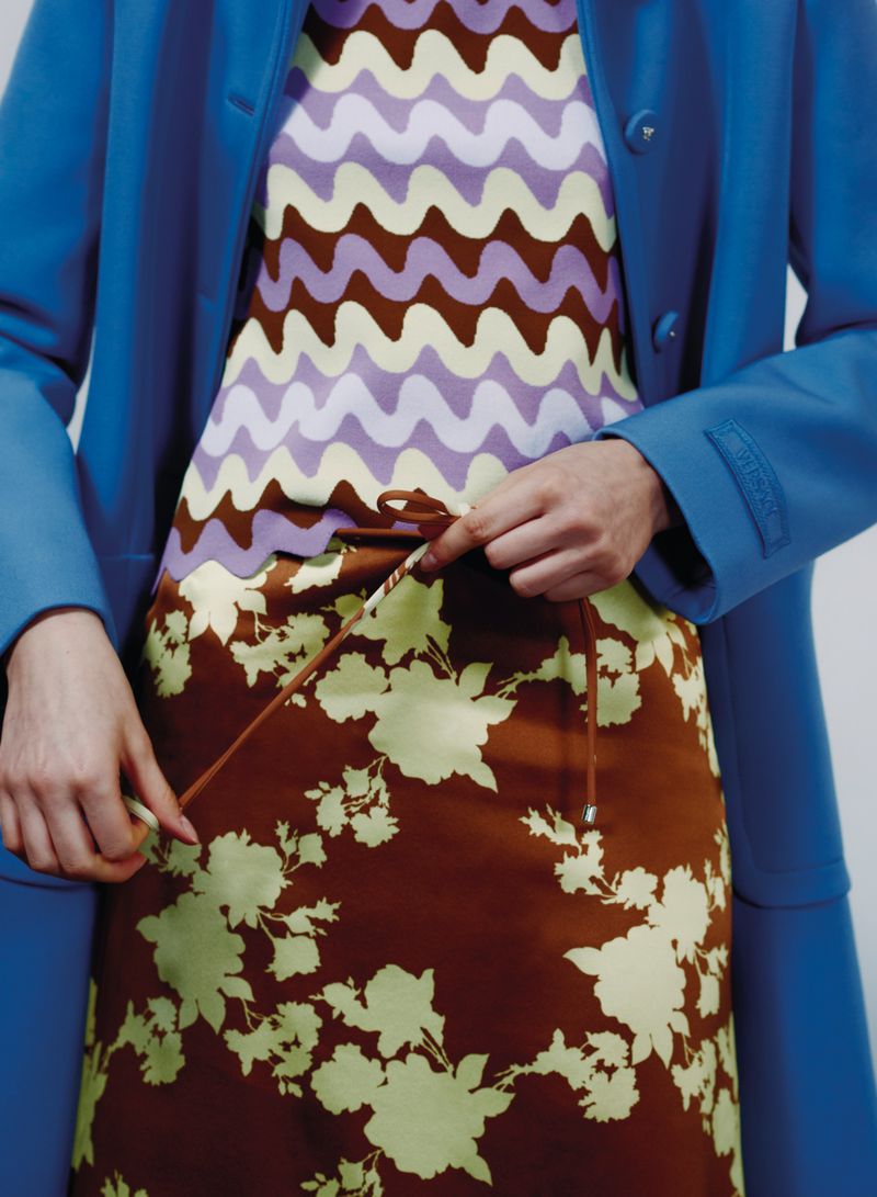 Close-up shot of woman adjusting the drawstring of a floral print skirt, paired with a patterned top and a bright blue coat indoors.