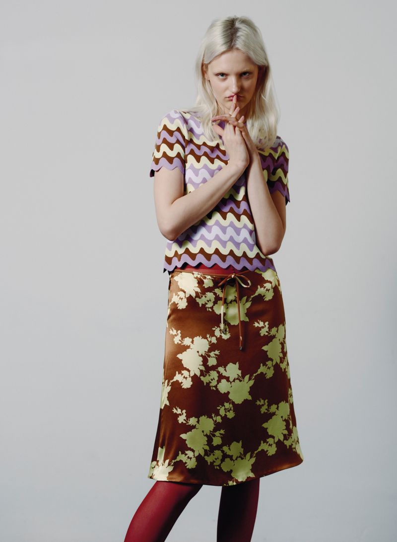 A woman with fair hair poses indoors, wearing a patterned top and floral print skirt. She is wearing red hosiery and faces forward.