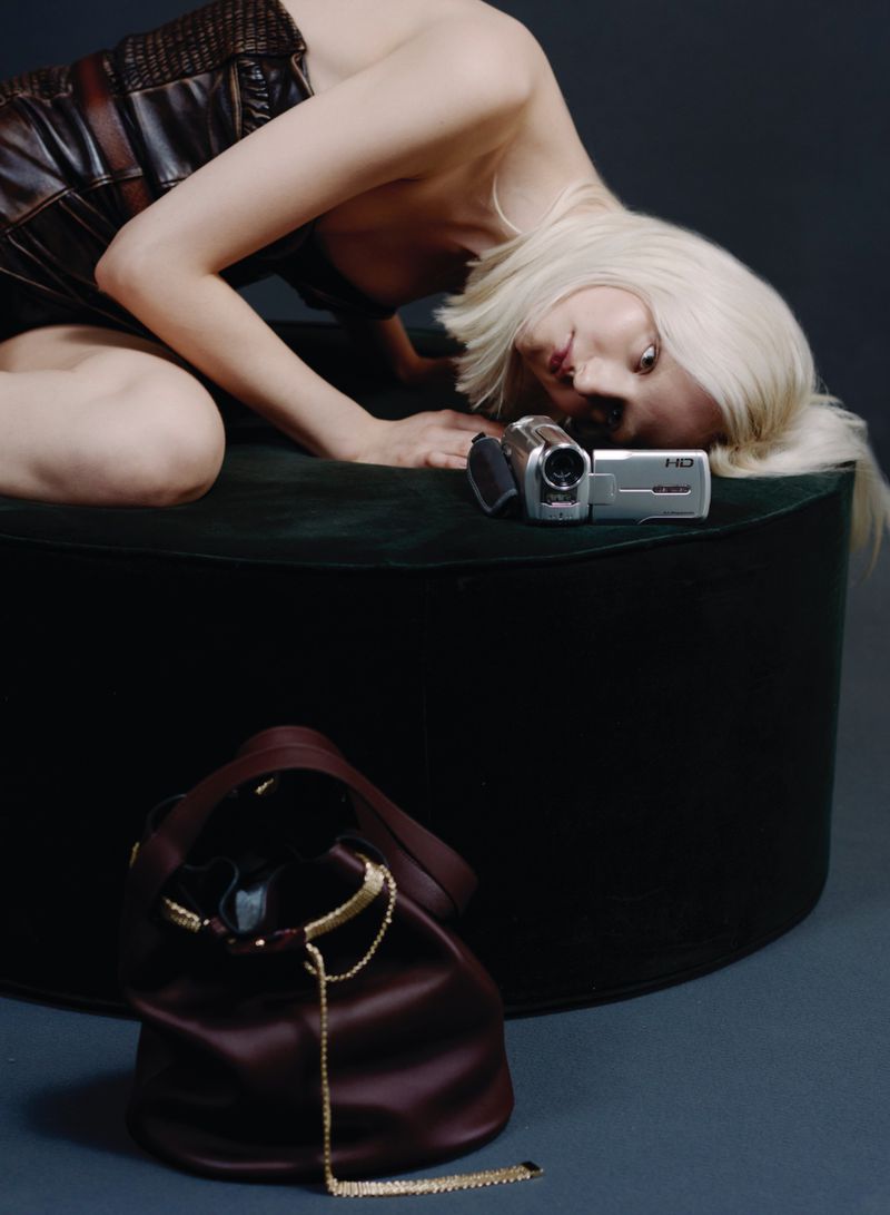 Woman in a black dress lying on a dark green cushion next to a vintage video camera with a burgundy leather drawstring bag.