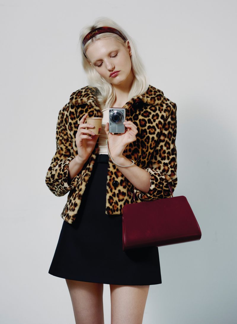 Young fair-skinned woman with blonde hair wearing a leopard-print jacket, black skirt, and headband, holds a camera indoors.