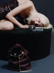 Model with blonde hair draped over a dark green stool alongside a silver camcorder and a dark burgundy leather bucket bag