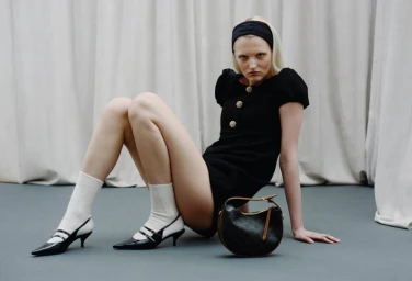 Model in black mini dress wearing white socks and heels, seated on a grey floor against a draped curtain background