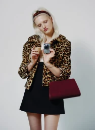 Model in leopard print faux fur jacket holding a small camera and a coffee cup against a plain white background