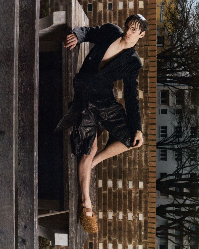 A young male model wears a dark pinstripe blazer and textured black shorts, leaning against a wooden bench in an outdoor urban setting.