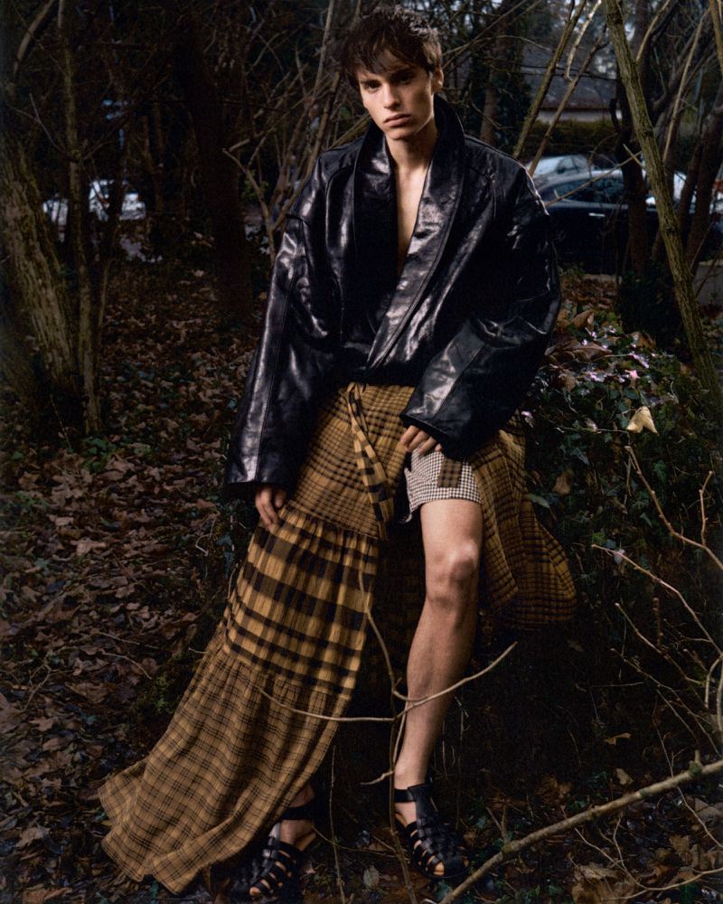 Fashionable young adult male poses outdoors wearing a black jacket, tan plaid skirt, and gladiator sandals among trees and foliage.