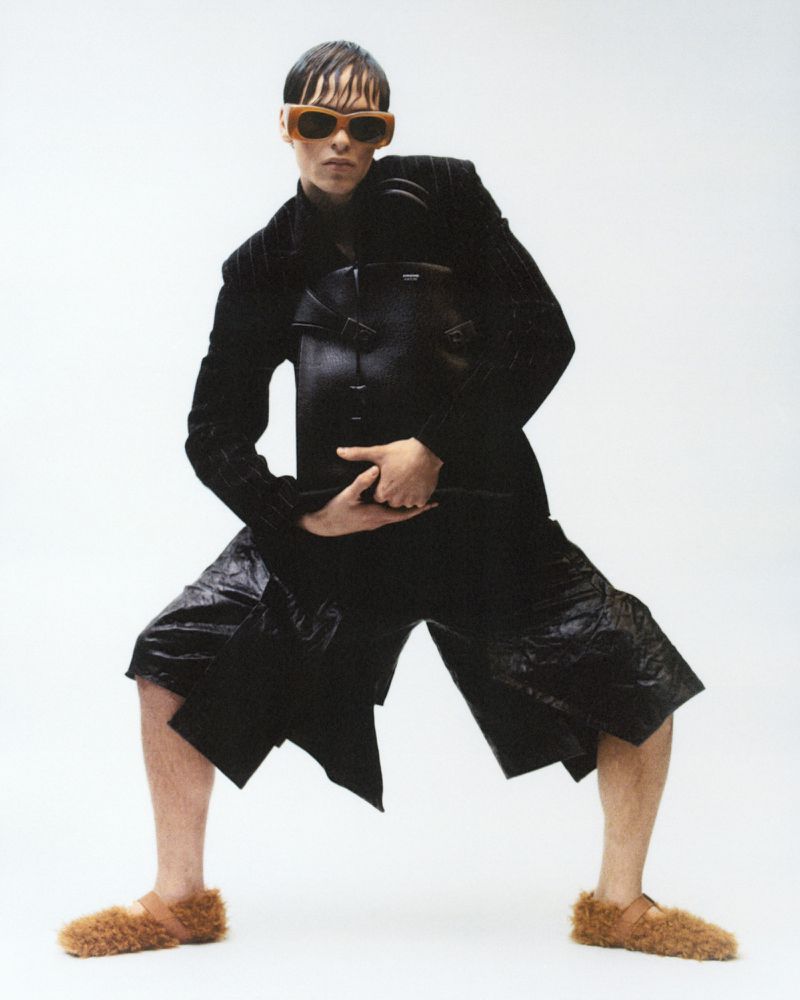 Stylish person in sunglasses and a black textured jacket and shorts strikes a pose in a fashion shot against a white backdrop.
