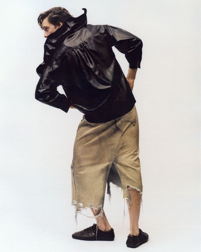 Man in black jacket poses in front of white background. Frayed beige skirt, black sneakers, and hands on hips complete fashion look.