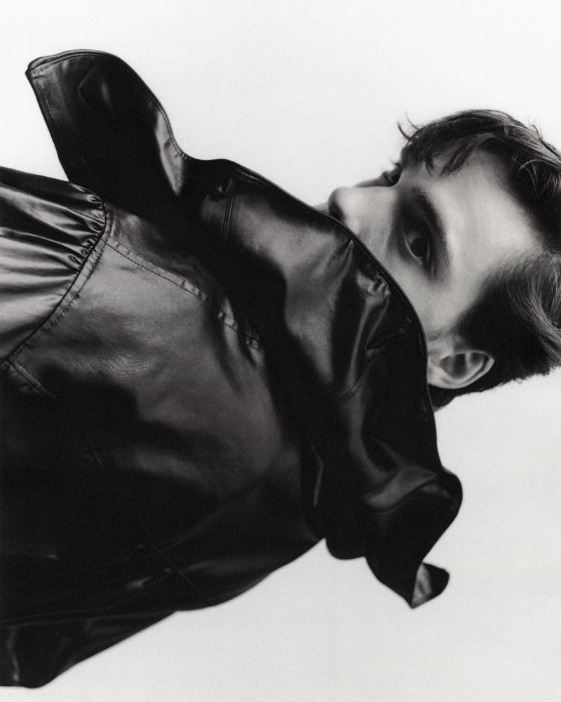 Portrait of a young adult man with dark hair, partially obscured by a shiny black jacket in a studio setting with a white backdrop.