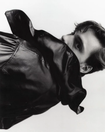 Portrait of a young adult man with dark hair, partially obscured by a shiny black jacket in a studio setting with a white backdrop.
