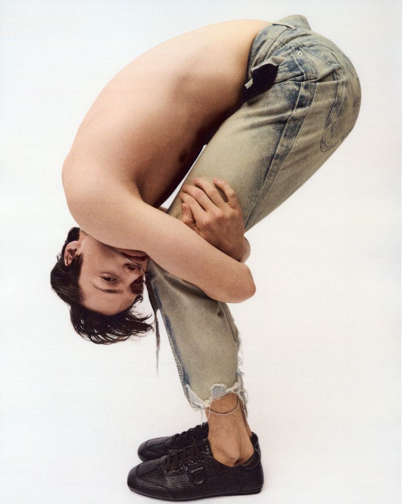 A slender adult man is bending over in a forward fold pose with his head near his knees, wearing jeans and black leather shoes on a white background.