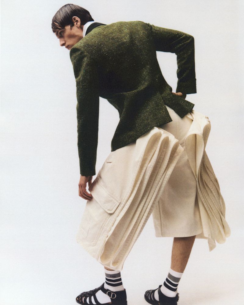 Fashionable adult male poses in a green tweed blazer, pleated ivory culottes, socks, and fisherman sandals in a brightly lit photo studio.