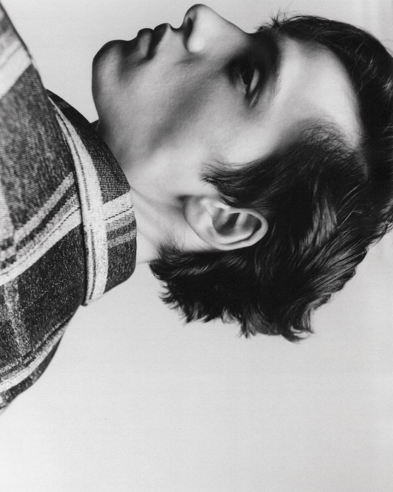 Monochrome portrait of a young man with dark hair wearing a plaid shirt, captured in a studio environment with soft, diffused lighting.