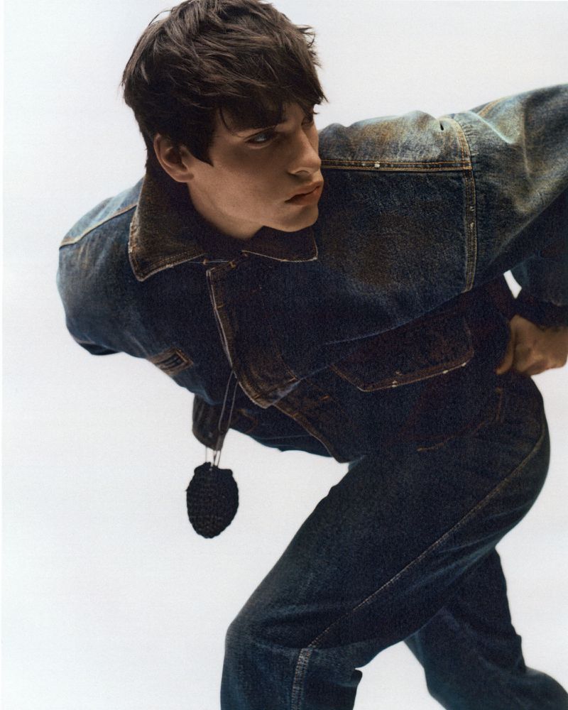 Dark-haired man in a denim jacket and jeans, standing against a white background, strikes a confident pose in a fashion-forward portrait.