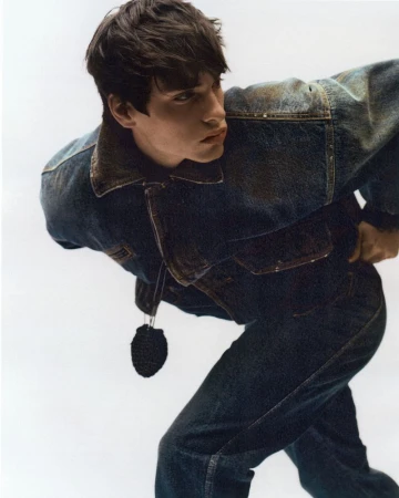 Young man in dark denim jacket leaning forward against a plain white studio background