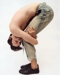 Shirtless man bending forward wearing denim jeans and black sneakers against a plain white background