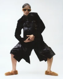 Model in a black jacket and shorts holding a large bag against a white background