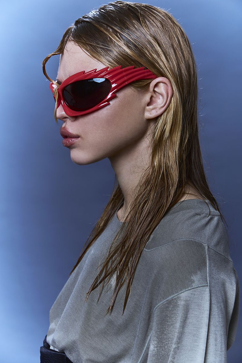 Woman in red sunglasses and grey top poses against a blue backdrop in a beauty and fashion editorial shot. Close-up studio shot.