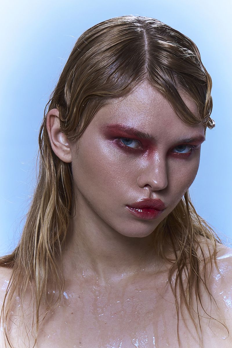 Close-up of a blonde woman with wet hair and red eyeshadow against a light blue background in a beauty and fashion context.