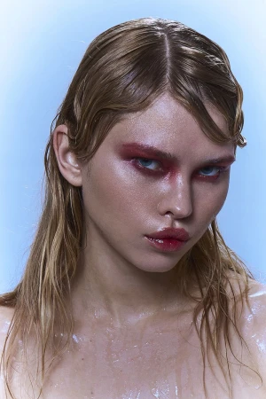 Close-up of a blonde woman with wet hair and red eyeshadow against a light blue background in a beauty and fashion context.