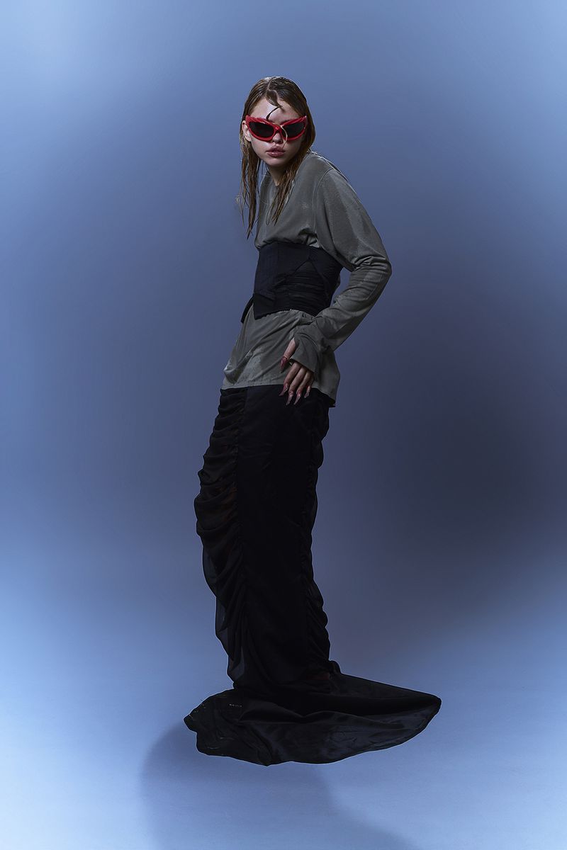 A young woman wearing red sunglasses, a grey top, a black corset and a long black skirt, poses in a studio with a blue background.