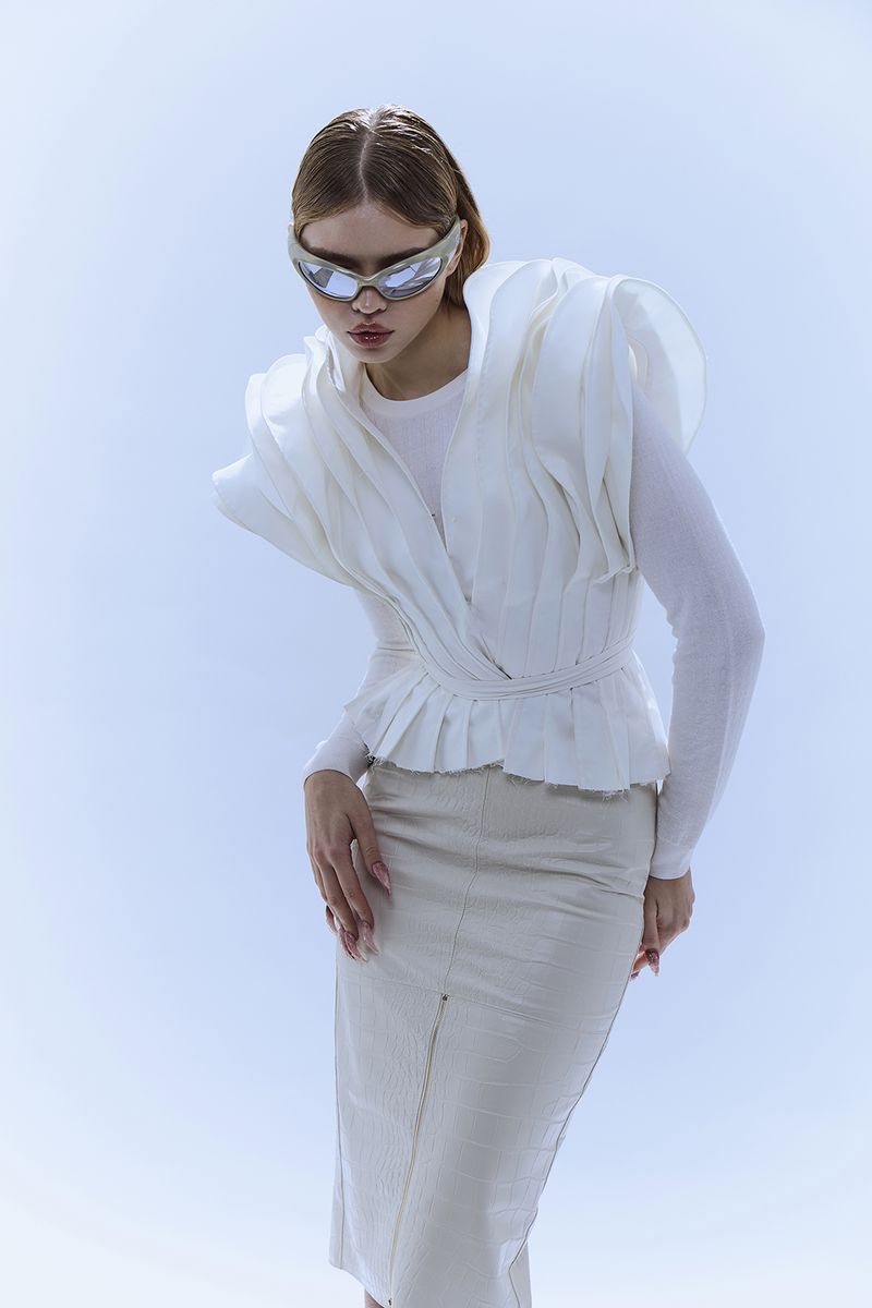 A woman in futuristic glasses poses in a bright studio wearing a white layered top and skirt, creating a modern fashion portrait.