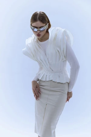 A woman in futuristic glasses poses in a bright studio wearing a white layered top and skirt, creating a modern fashion portrait.