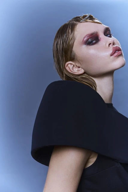 Headshot of a young woman with bold, red eye makeup and slicked-back hair against a light blue background, wearing a black top.