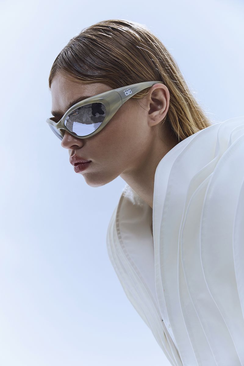 A woman with light brown hair is wearing fashionable sunglasses and a white jacket against a bright blue and white background.