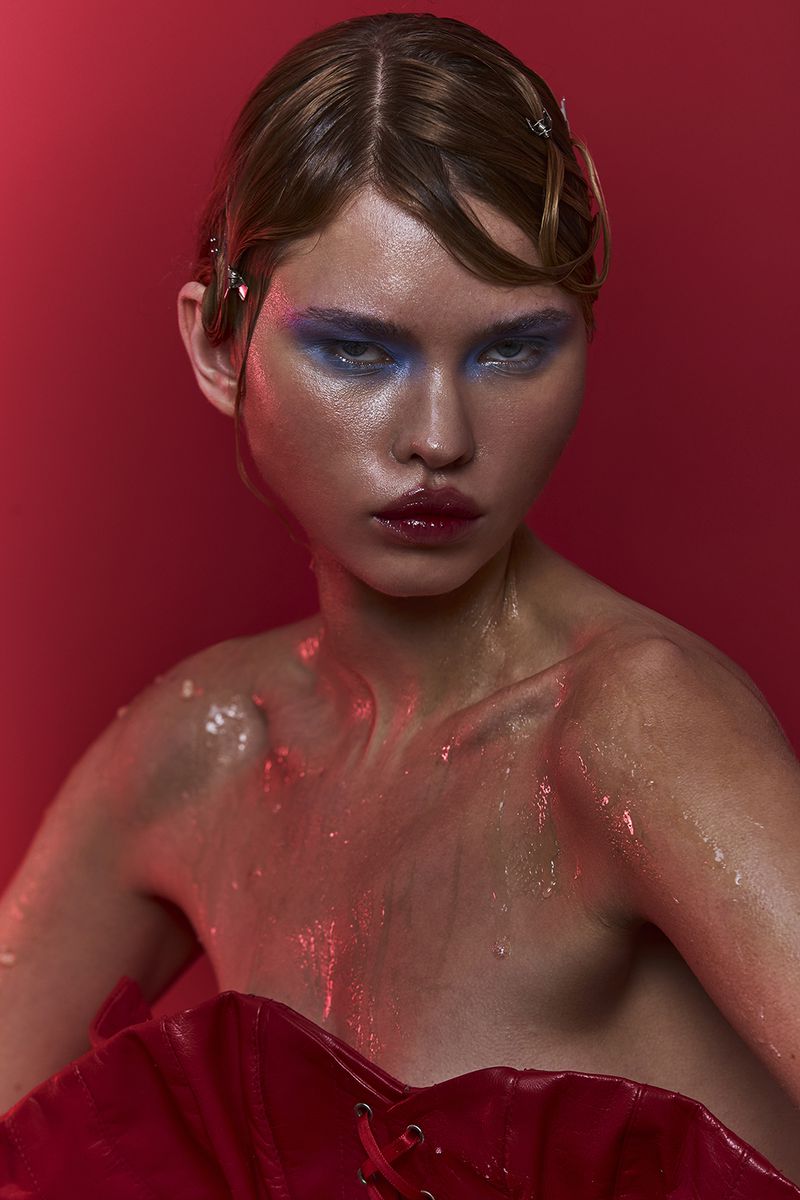 Close-up shot of a woman with bronze skin, blue eyeshadow, and glossy red lips against a vibrant red backdrop, glossy skin and hair.