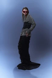 Model wearing red sunglasses, olive long-sleeve top, black corset and black skirt against blue background for PAP Magazine project Arkhé