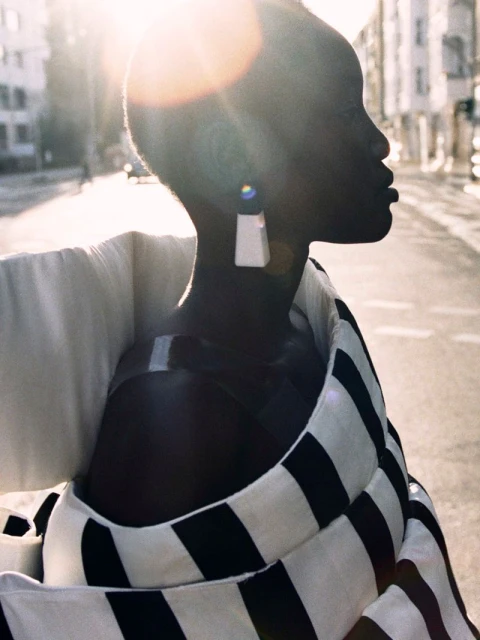 Elegant black woman in a striped jacket, posing on a city street under bright sunlight, wearing statement geometric earrings.