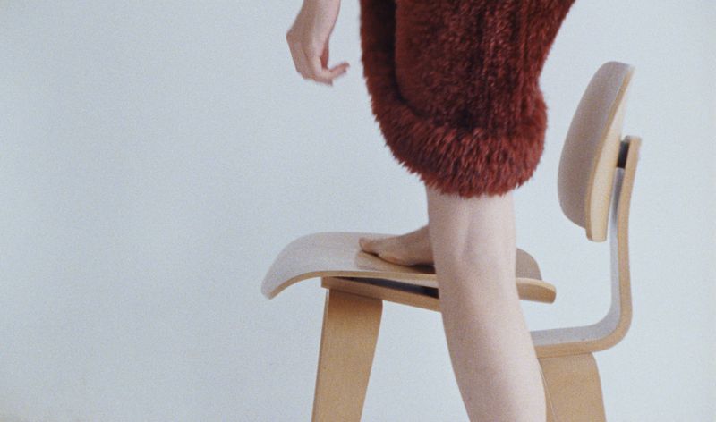 Fuzzy red dress garment detail on light wood chair in white studio. Seated human figure with bare legs. Fashion shot with soft lighting.
