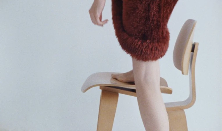 Fuzzy red dress garment detail on light wood chair in white studio. Seated human figure with bare legs. Fashion shot with soft lighting.