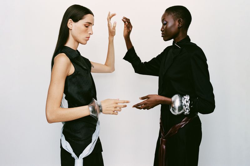 Two women face each other in a fashion shot. One wears a black sleeveless top with white accents and the other a black jacket with a brown belt.