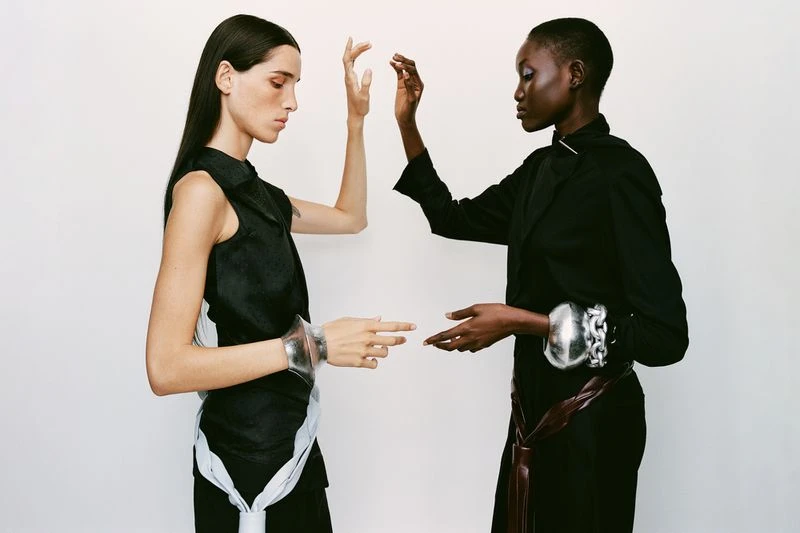 Two women face each other in a fashion shot. One wears a black sleeveless top with white accents and the other a black jacket with a brown belt.