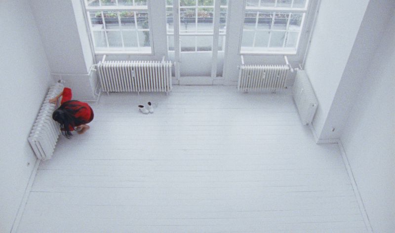 High-angle view of a person kneeling in an all-white room with radiators and windows, creating a minimalist and bright visual.