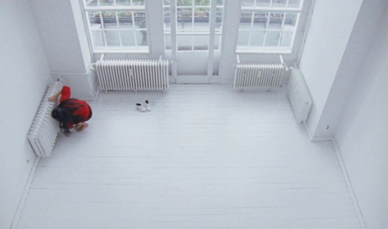 High-angle view of a person kneeling in an all-white room with radiators and windows, creating a minimalist and bright visual.