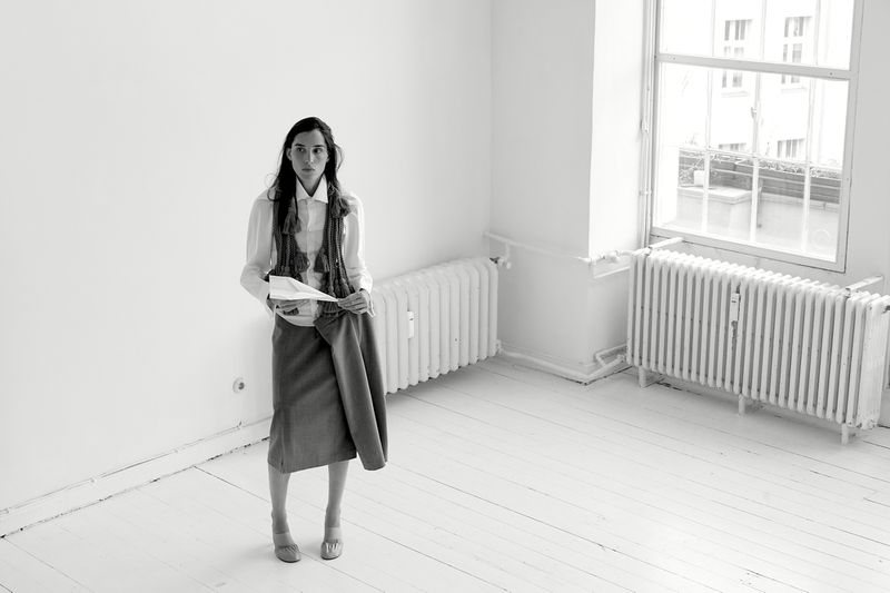 Monochrome portrait of a stylish woman in a white shirt, vest, skirt, and heels, holding a skirt in a minimalist interior space.