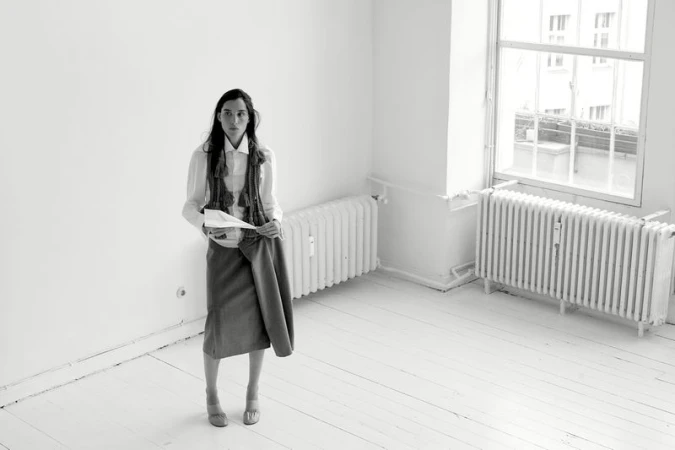Monochrome portrait of a stylish woman in a white shirt, vest, skirt, and heels, holding a skirt in a minimalist interior space.