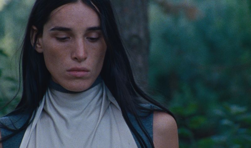 Close-up of a woman with dark hair in a forest. The lighting is soft, and the subject wears a light-colored high-necked garment.