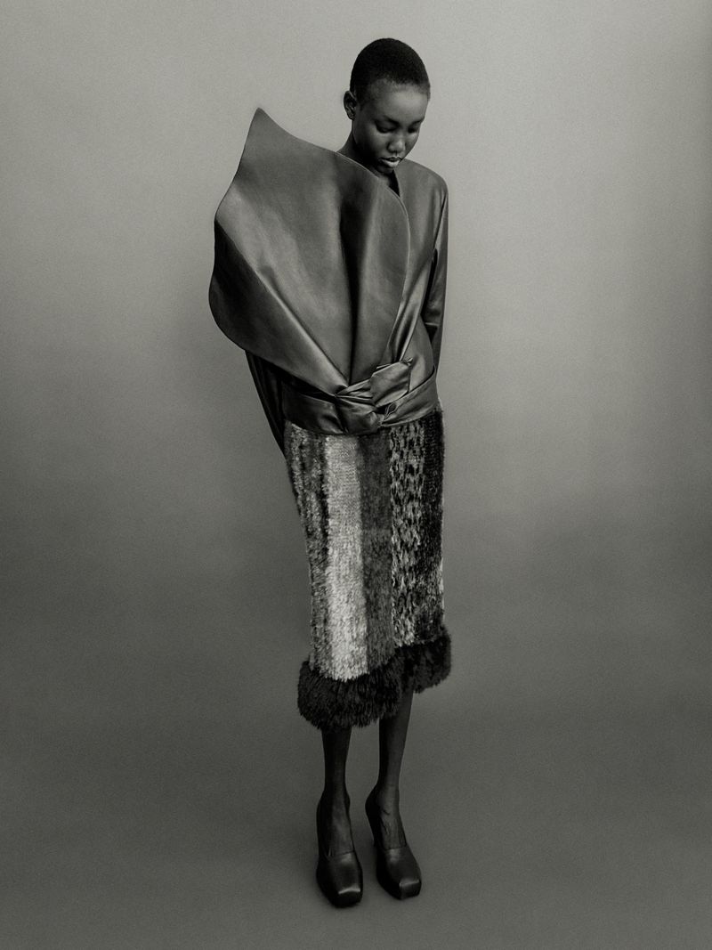 Black and white fashion portrait of a slender model in a voluminous top and patterned skirt, in a studio setting, looking downward.