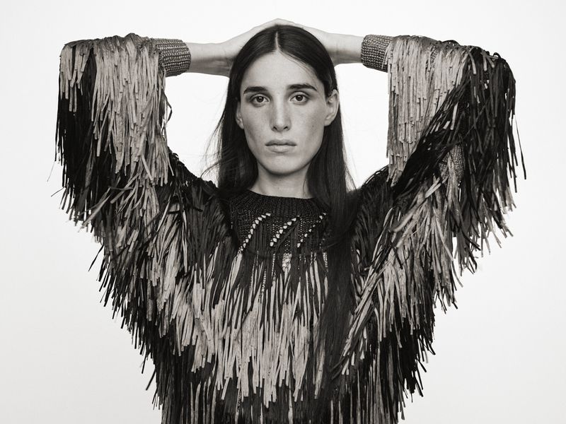 Black and white portrait of a woman in a dark top with light and dark fringe sleeves, hands behind her head, against a white backdrop.