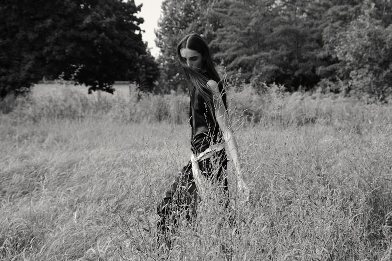 Black and white portrait of a young woman in black top walking through tall grassy field surrounded by trees and vegetation.