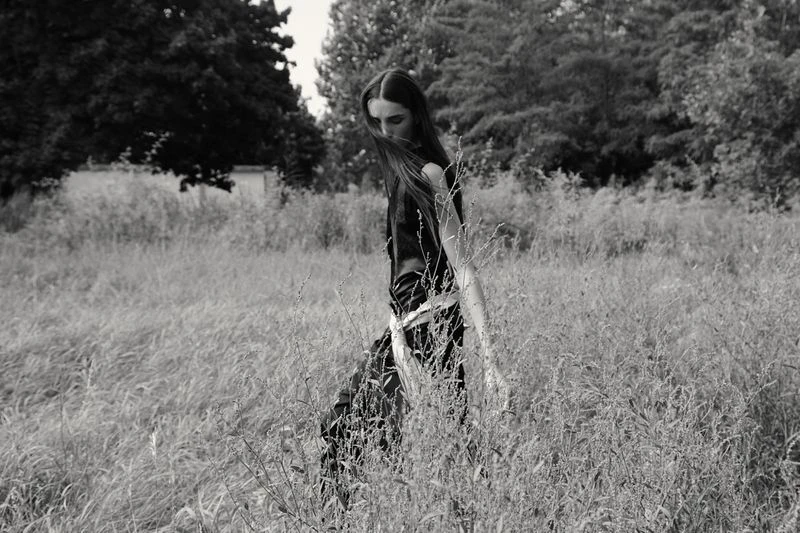 Black and white portrait of a young woman in black top walking through tall grassy field surrounded by trees and vegetation.