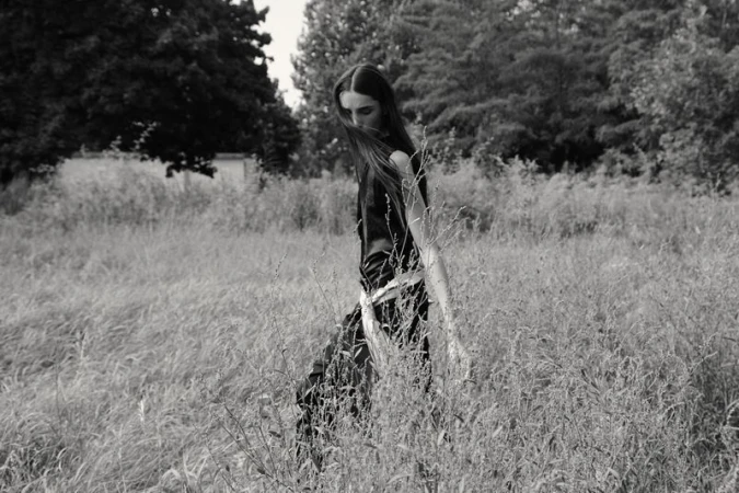 Black and white portrait of a young woman in black top walking through tall grassy field surrounded by trees and vegetation.