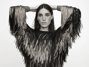 Model with long dark hair wearing a black fringed textured sweater with hands behind head against a white background