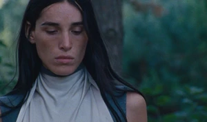 Close-up portrait of a woman with long dark hair wearing a high-neck grey and white top outdoors against a blurred green forest backdrop.