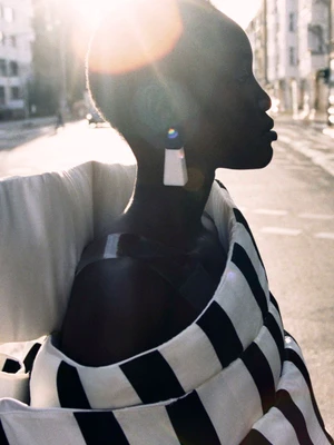 Black woman in profile wearing a black and white striped garment and statement earring on a sunlit street.